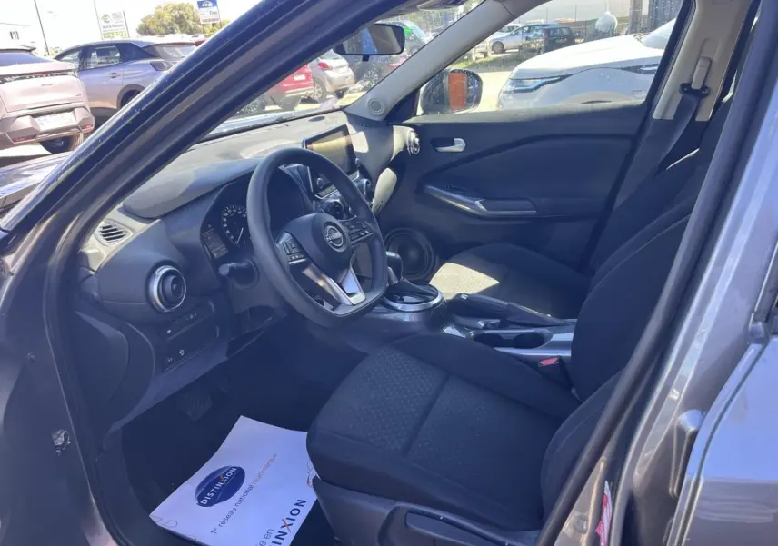Vue intérieure côté conducteur du Nissan Juke 2023 gris argile, montrant le volant multifonction et les sièges en tissu noir.