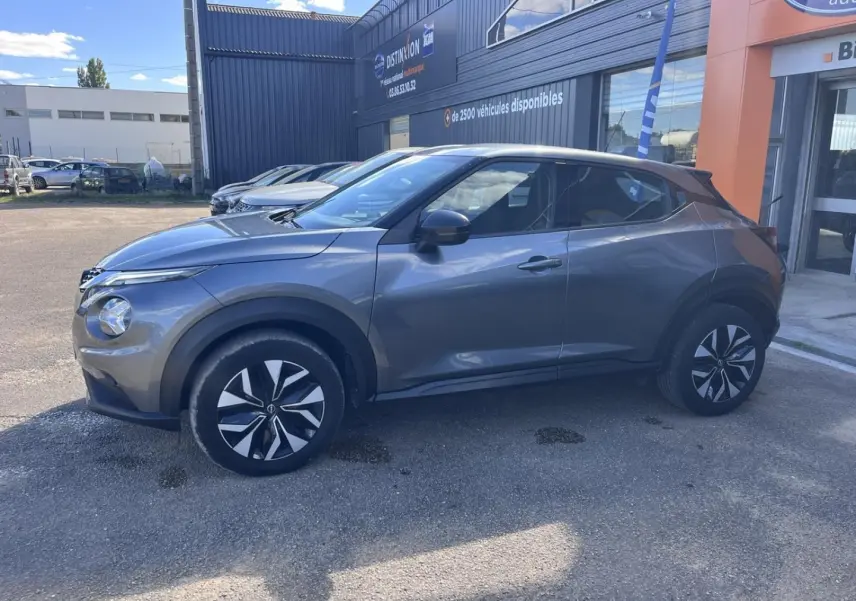 Profil droit d'un Nissan Juke 2023 gris argile avec jantes bi-ton et poignées ton carrosserie, stationné devant un bâtiment.