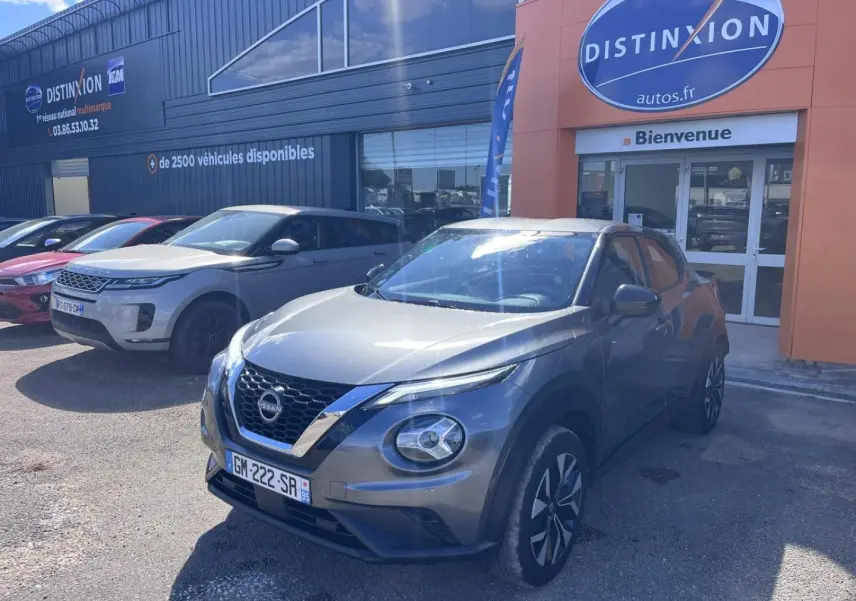 Nissan Juke gris argile vue 3/4 avant droit, avec jantes bi-ton et calandre chromée, stationné devant un concessionnaire.