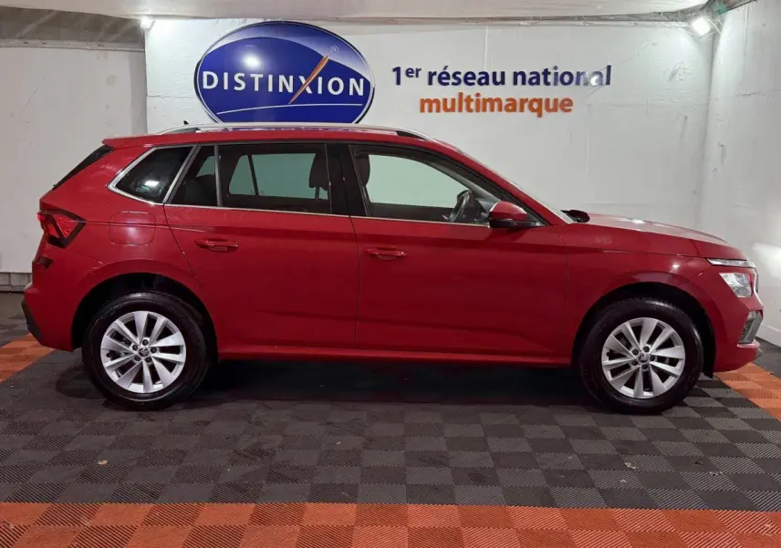 Vue latérale droite d’un Skoda Kamiq rouge 2024 avec jantes alliage et barres de toit noires en intérieur showroom.