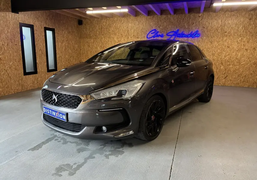 DS5 gris 3/4 avant droit en intérieur, avec jantes noires et calandre distinctive DS sous néon bleu.