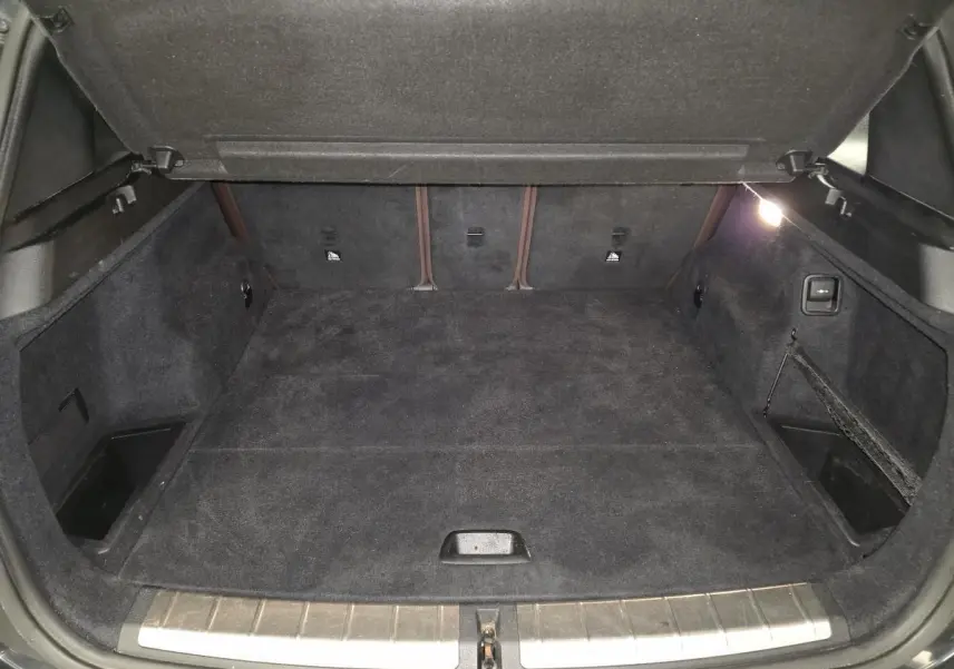 Coffre ouvert vu de face, intérieur noir avec tapis et rangements latéraux, hayon électrique visible sur BMW X1 noir.