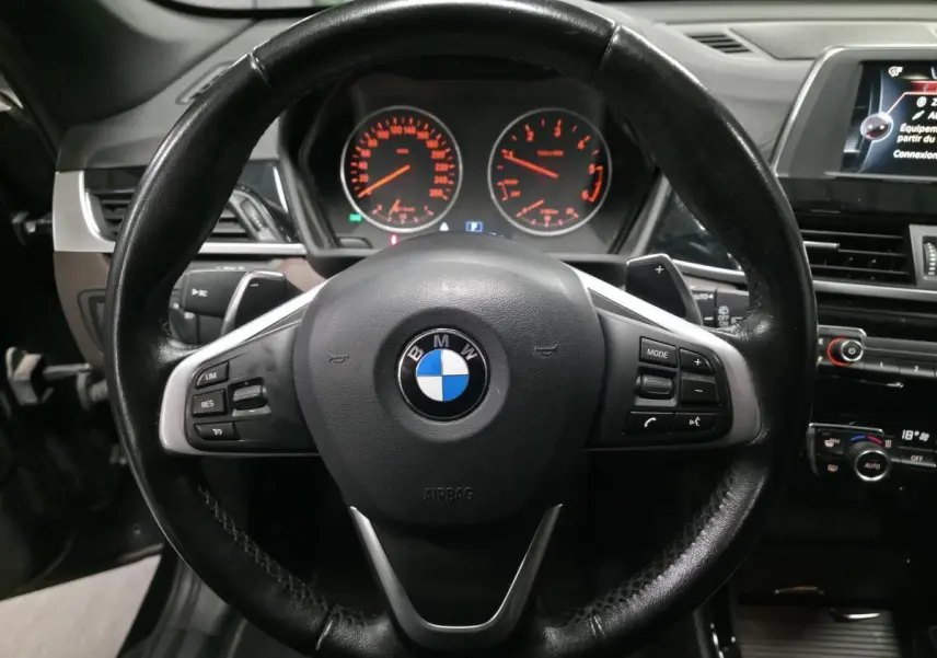 Vue rapprochée du volant cuir noir multifonctions d'une BMW X1 xDrive 25d 2016 avec tableau de bord et écran central visibles.