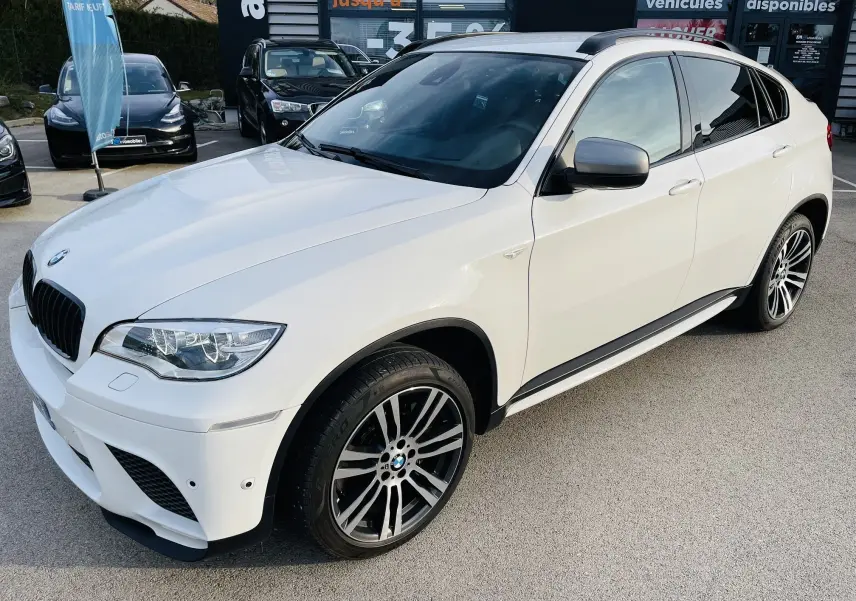 BMW X6 blanc en 3/4 avant droit, avec jantes sport et calandre caractéristique M, stationné en extérieur.