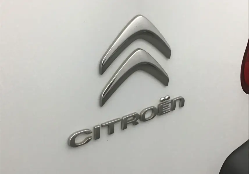 Gros plan sur le logo argenté Citroën sur la porte arrière blanche d'un Berlingo 2018, avec un feu arrière partiellement visible.