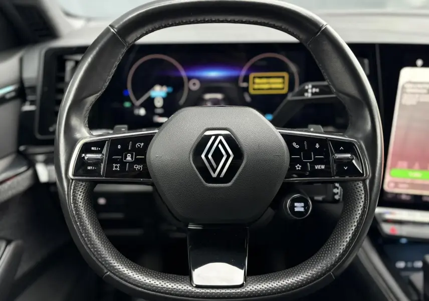 Vue rapprochée du volant noir perforé avec logo Renault au centre, tableau de bord numérique en arrière-plan flou.