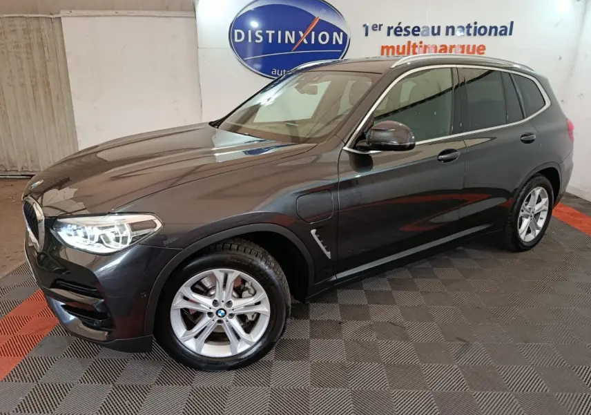BMW X3 xDrive30e gris foncé vue 3/4 avant droit avec phares allumés dans un showroom intérieur.
