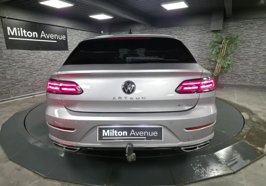 Vue arrière d'un Volkswagen Arteon gris avec feux LED allumés et dispositif d'attelage rabattable visible en intérieur showroom.