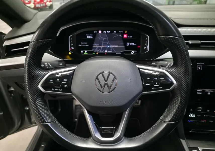 Vue frontale du volant noir perforé avec logo Volkswagen et tableau de bord digital affichant la navigation du Volkswagen Arteon Shooting Brake gris.