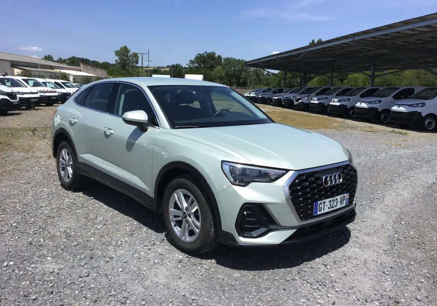 Audi Q3 Sportback gris clair métal vue 3/4 avant droit sur un terrain gravillonné avec plusieurs véhicules blancs en arrière-plan.