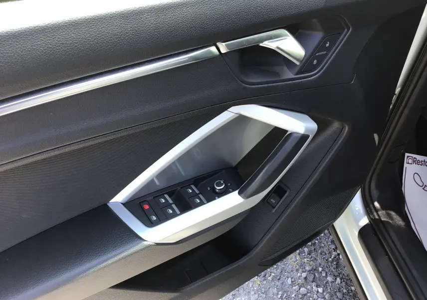 Vue rapprochée de la porte avant gauche de l'Audi Q3 Sportback gris clair, avec commandes électriques et poignée argentée.