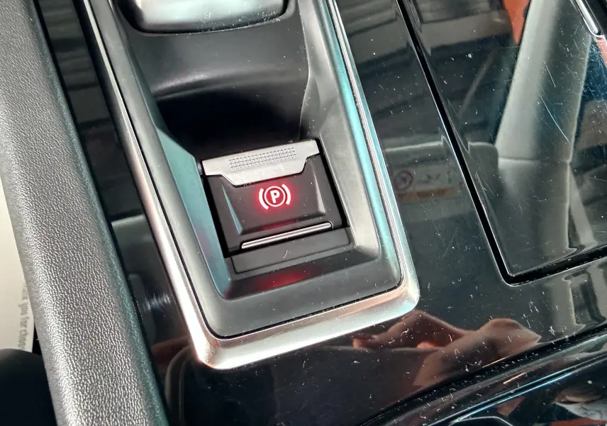 Gros plan sur le bouton de frein à main électrique rouge dans la console centrale noire brillante d'une Peugeot 308 SW noire.