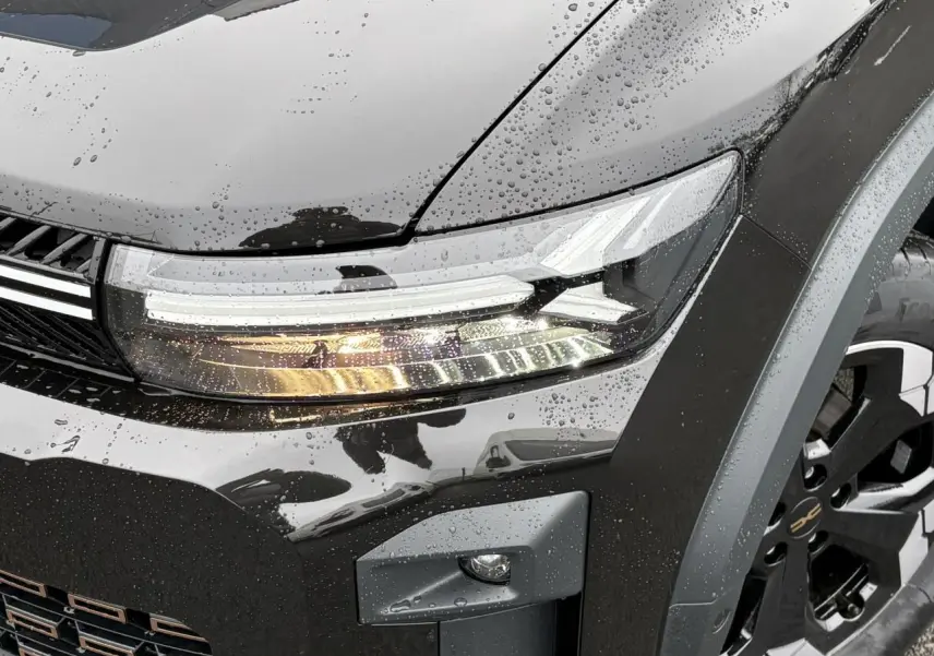 Gros plan sur le phare avant droit et la jante noire diamantée du Dacia Bigster noir nacré sous la pluie.