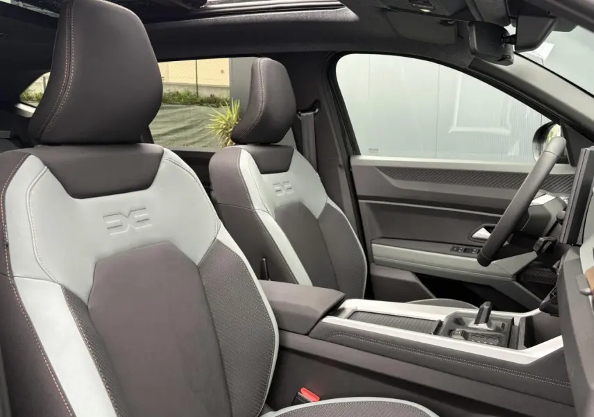 Vue intérieure côté droit du Dacia Bigster 2026, sièges bicolores gris clair et foncé avec logo embossé, toit panoramique visible.