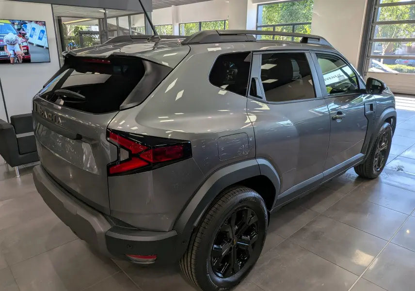 Vue 3/4 arrière droit du Dacia Duster 2026 gris schiste avec jantes noires et barres de toit en showroom.