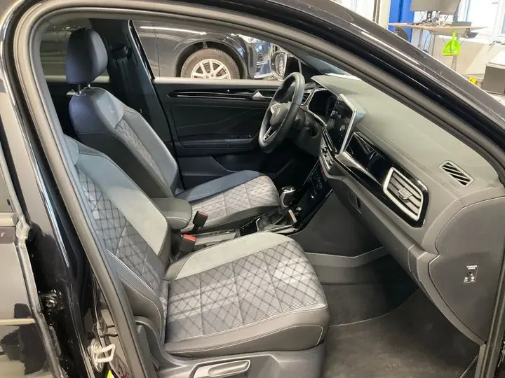 Vue intérieure côté conducteur du Volkswagen T-Roc noir intense 2025, sièges gris à motif losange et tableau de bord moderne.