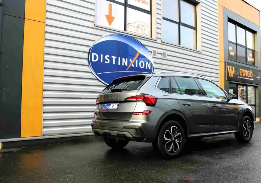 Vue 3/4 arrière droite d'un Skoda Kamiq gris graphite 2025 garé devant un bâtiment commercial.