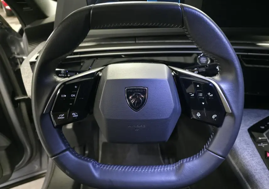 Volant cuir noir avec logo Peugeot en vue frontale, tableau de bord et console centrale tissu gris visibles.