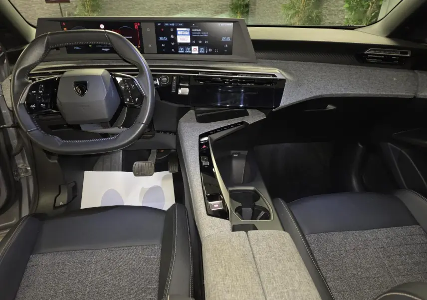 Vue intérieure avant du Peugeot 3008 Hybrid 2025 avec tableau de bord tissu gris, volant compact cuir noir et double écran HD flottant.