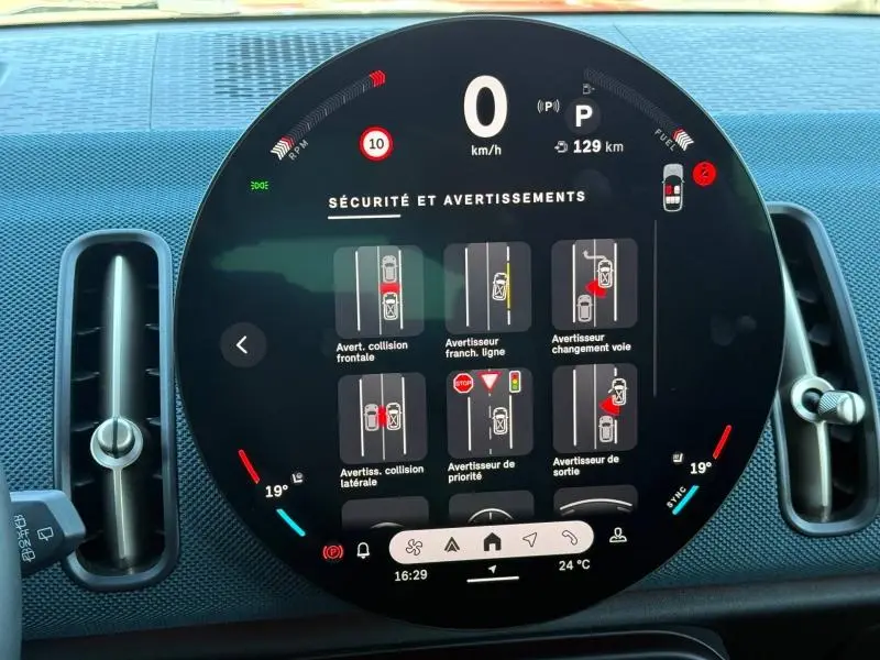 Vue rapprochée du tableau de bord numérique circulaire du MINI Countryman 2024 affichant les alertes de sécurité, entouré d'aérateurs verticaux.