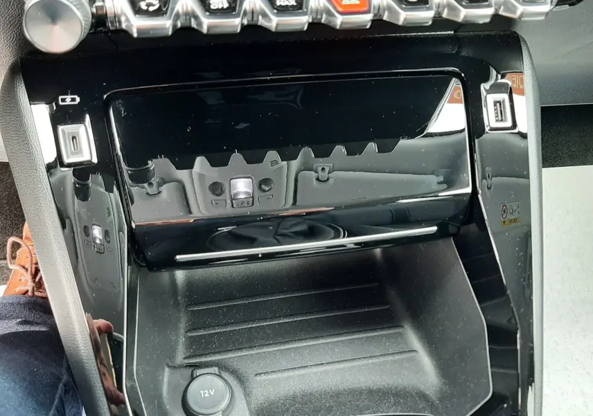 Vue rapprochée de la console centrale noire brillante du Peugeot 2008 1.5 BlueHDi 110 Allure avec commandes et prise 12V visibles.