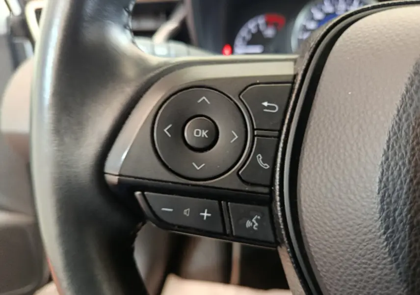 Gros plan sur les commandes gauche du volant cuir noir de la Toyota Corolla Hybride 122h Active, avec tableau de bord flou en arrière-plan.