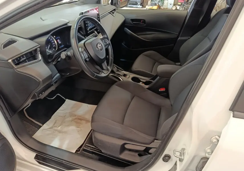 Vue intérieure côté conducteur de la Toyota Corolla Hybride blanche 2020, avec tableau de bord et sièges noirs.