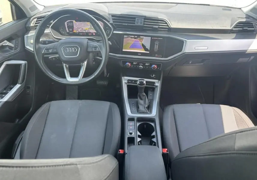 Vue intérieure avant de l'Audi Q3 blanc, montrant le volant multifonctions, écran central avec caméra de recul et sellerie tissu noire.