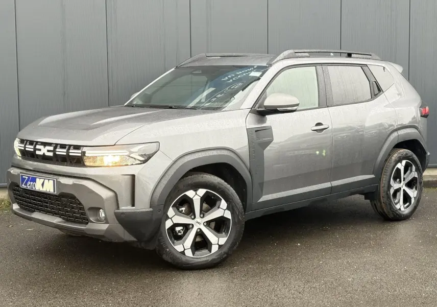 Dacia Duster gris schiste en 3/4 avant droit, avec jantes alliage diamantées et barres de toit noires.