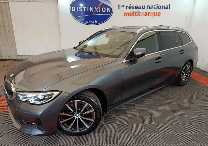 BMW Série 3 318d gris clair vue 3/4 avant droit, avec jantes alliage et phares LED allumés.