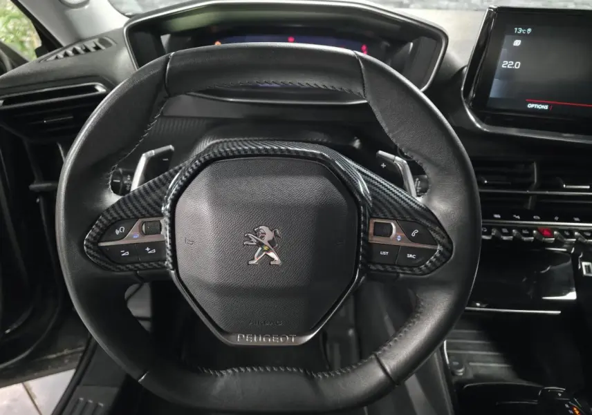 Volant compact cuir noir avec insert carbone et logo Peugeot, vue rapprochée du tableau de bord et écran tactile.