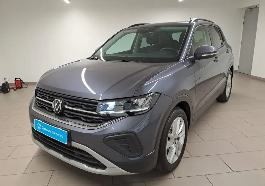 Volkswagen T-Cross gris cendré en 3/4 avant droit, avec calandre noire et phares allumés en intérieur showroom.