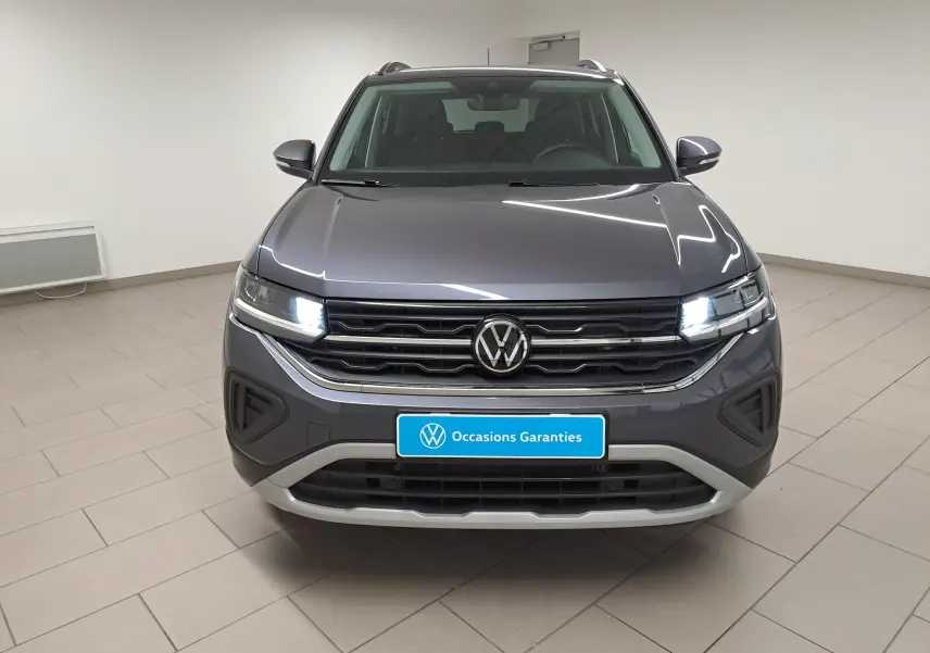Vue frontale du Volkswagen T-Cross 2025 gris cendré métallisé avec phares allumés dans un showroom.