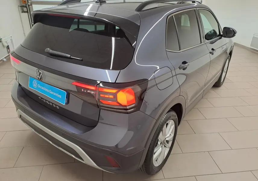 Vue 3/4 arrière droit du Volkswagen T-Cross 2025 gris cendré avec feux arrière allumés et toit avec barres de toit.