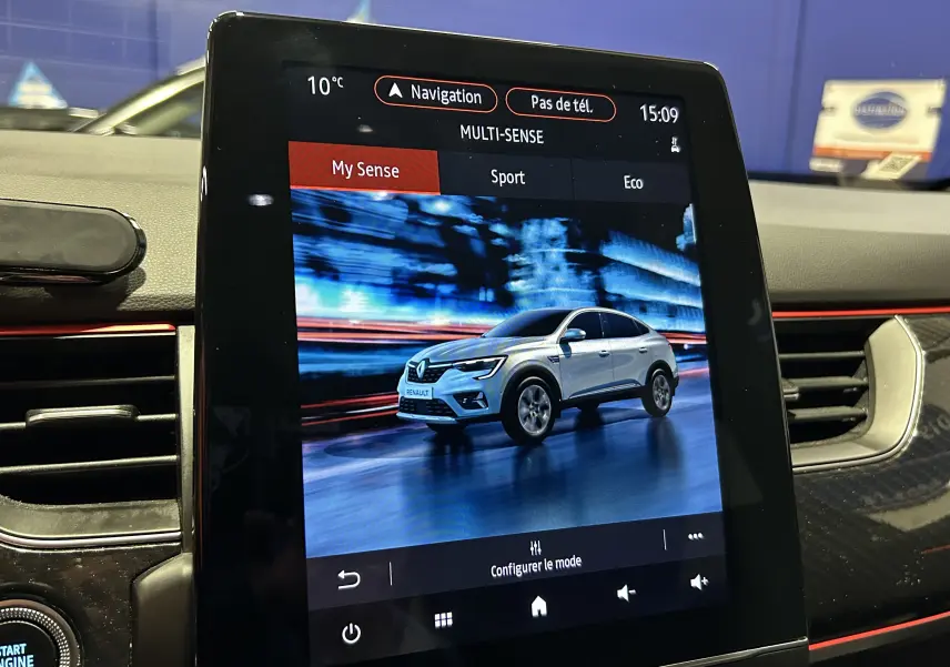Écran tactile intérieur montrant un Renault Arkana blanc en mouvement sur fond urbain flou, mode My Sense activé.