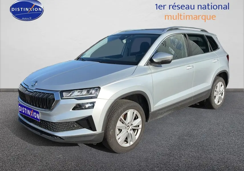 SUV Skoda Karoq gris en 3/4 avant droit avec calandre noire et jantes alu visibles sur sol goudronné.