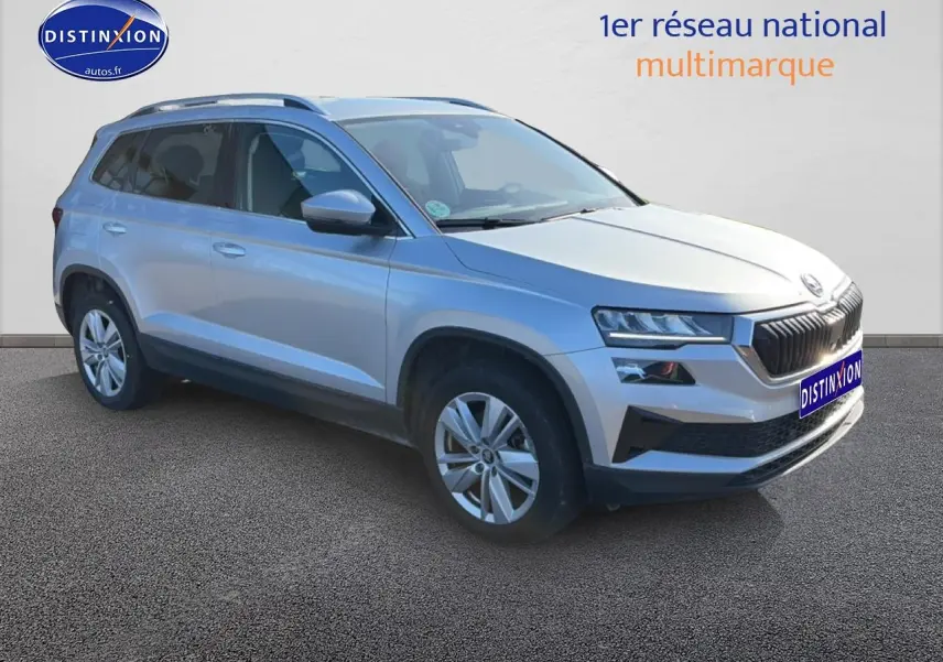 SKODA KAROQ gris 3/4 avant droit, jantes alu, barres de toit et feux LED visibles sur fond neutre.