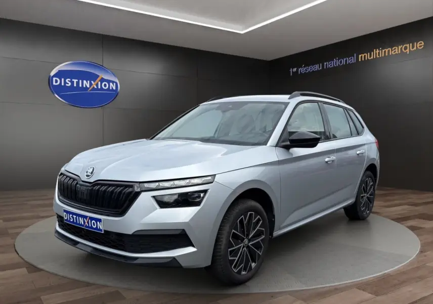 Vue 3/4 avant d'un Skoda Kamiq gris clair avec jantes bi-ton et rétroviseurs noirs dans un showroom.