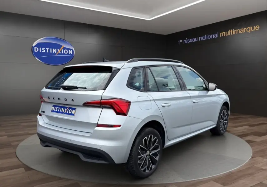 Vue 3/4 arrière droite d’un Skoda Kamiq gris clair avec jantes bi-ton et barres de toit noires.