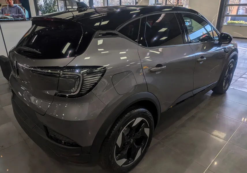 Renault Captur 2025 gris Cassiopé toit noir, vue 3/4 arrière droit en intérieur showroom, jantes biton 18 pouces.