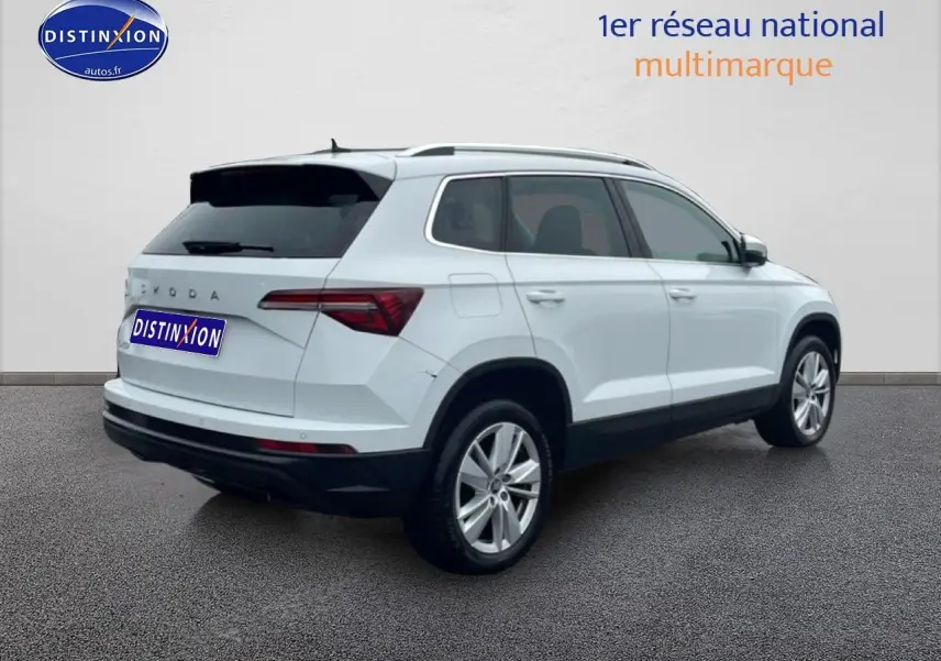 Vue 3/4 arrière droite d'un Skoda Karoq blanc glacial avec jantes alu et barres de toit visibles.