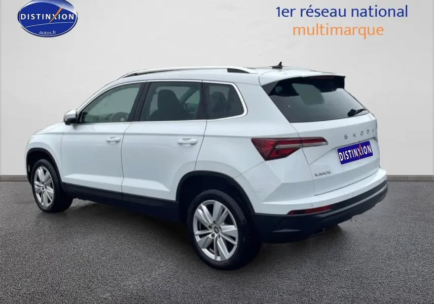 Vue 3/4 arrière droite d'un Skoda Karoq blanc glacial avec jantes alu et barres de toit sur fond neutre.