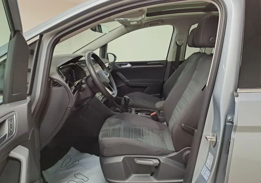 Vue intérieure côté conducteur d'un Volkswagen Touran gris argent 2025, sièges tissu noirs et tableau de bord moderne.