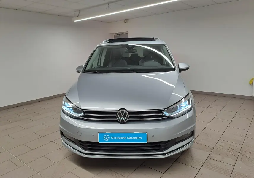 Volkswagen Touran gris argent vu de face, avec phares allumés et toit ouvrant partiellement ouvert.