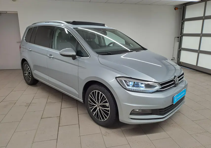 Vue 3/4 avant droit d'un Volkswagen Touran gris argent avec toit ouvrant panoramique en intérieur showroom.