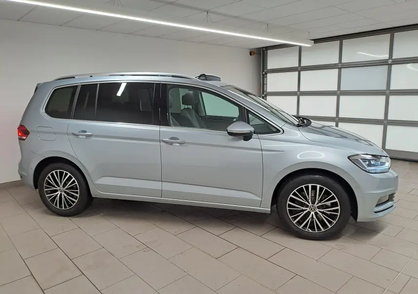 Vue de profil côté gauche d’un Volkswagen Touran gris argent avec jantes alliage et toit ouvrant panoramique en intérieur showroom.