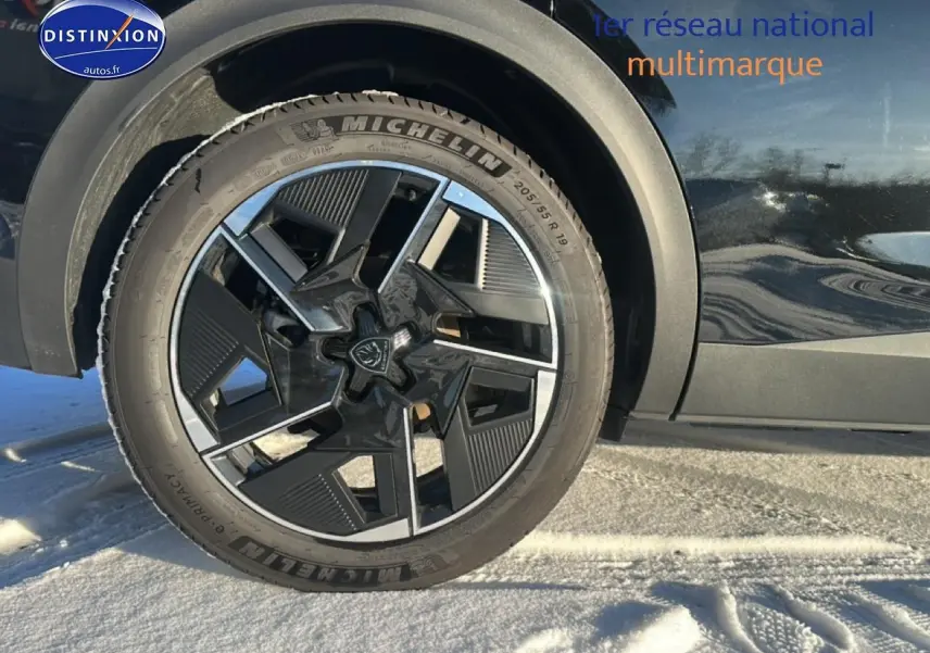 Gros plan sur la roue avant gauche et la jante alu noire du Peugeot 408 Hybrid 145 GT e-dcs6 noir sur sol enneigé.