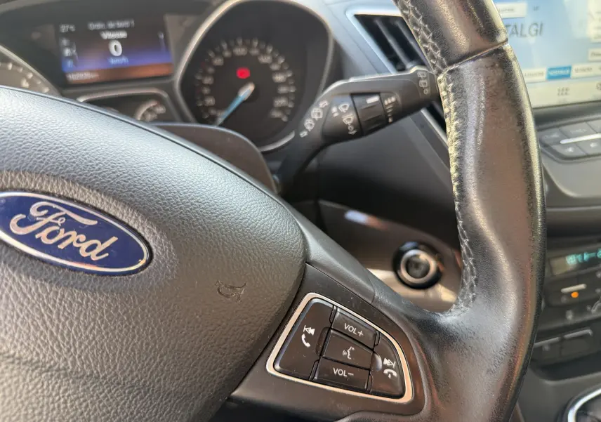 Vue rapprochée du volant noir de la Ford C-MAX 2018, montrant les commandes audio et le tableau de bord flou en arrière-plan.