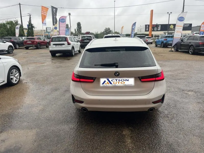 Vue arrière d'une BMW Série 3 316dA Business Design en blanc Alpinweiss avec feux arrière LED allumés sur parking humide.