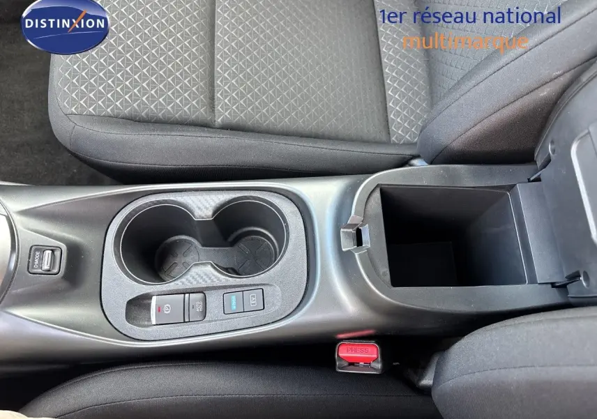 Vue plongeante sur la console centrale du Nissan Juke Hybrid 2024, avec porte-gobelets et rangement ouvert, sièges tissu noir.